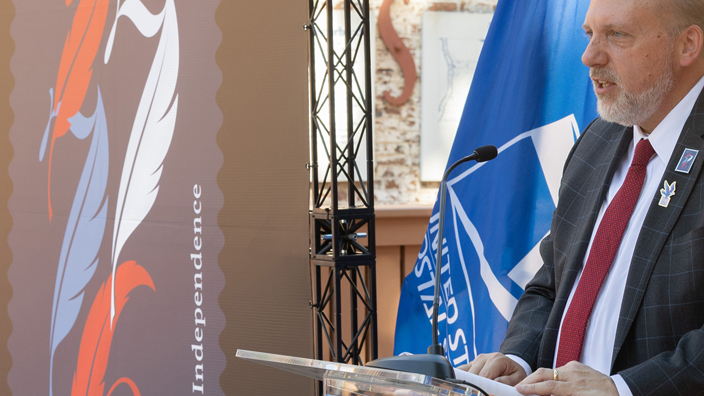 A man in a dark suit speaks from behind a lectern with a large image of the Declaration of Independence stamp off to his side.