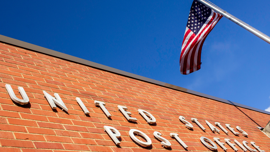 Brick exterior of a Post Office building with an American flag blowing in the wind out front