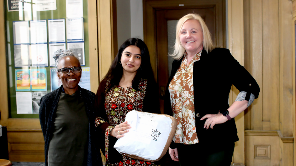 Three women standing on either side of a woman in the center who is holding a package