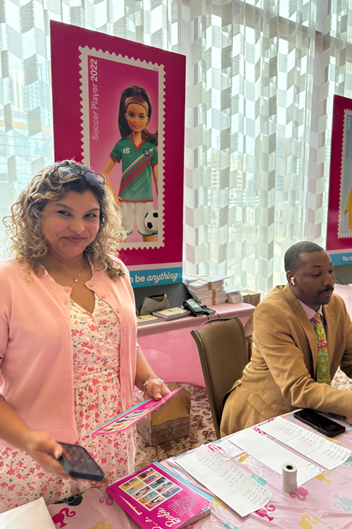 A standing woman wearing a pink dress and sitting man wearing a pink long-sleeve shirt under a beige jacket behind a table.