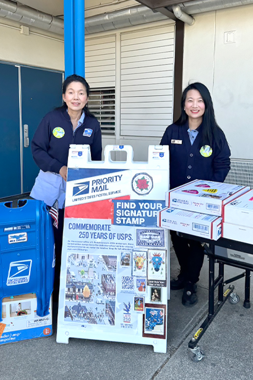 Two USPS employees standing behind a sandwich board display and table promoting Postal Service products