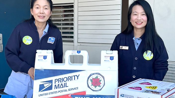 Two USPS employees standing behind a sandwich board display and table promoting Postal Service products