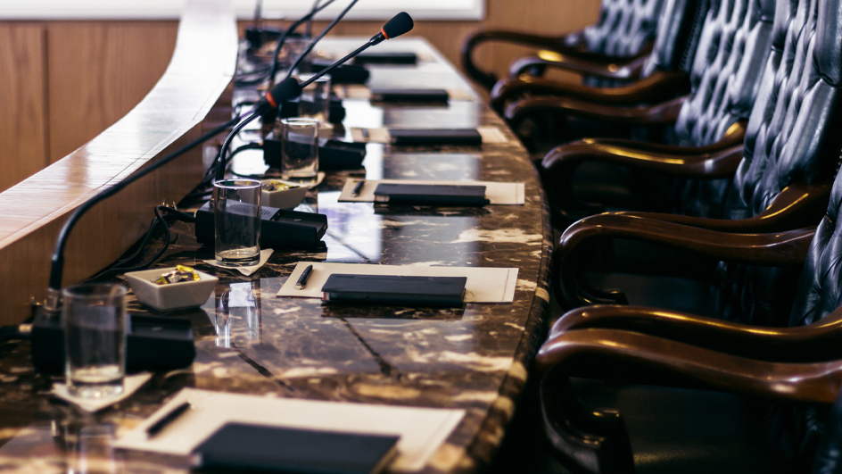 An image of high-back leather chairs behind a curved table containing microphones.