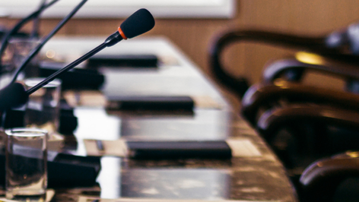 An image of high-back leather chairs behind a curved table containing microphones.