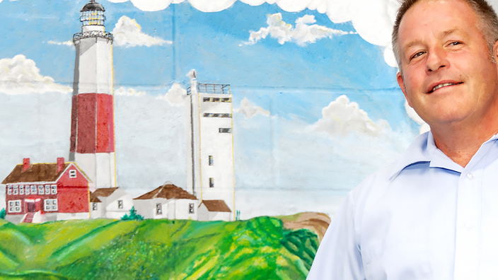USPS employee in light blue shirt standing to the right of a mural of lighthouse painted on a wall