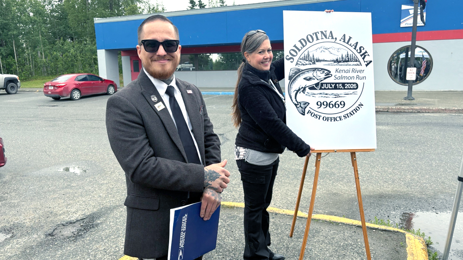 A man in a dark suit and sunglasses and a woman wearing black smile while standing beside an enlarged image of a pictorial postmark