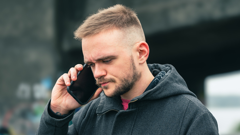 Man wearing a black coat and standing outdoors while holding a cellphone to his ear