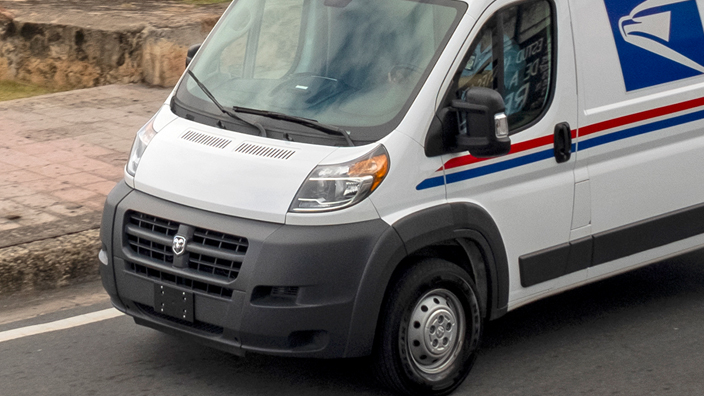 USPS delivery van driving on a road parallel to a large body of water