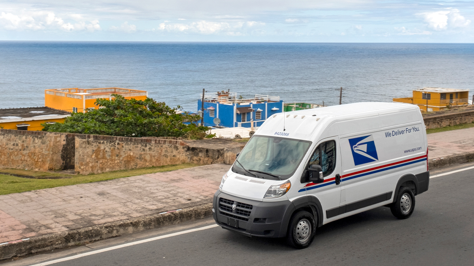 USPS delivery van driving on a road parallel to a large body of water