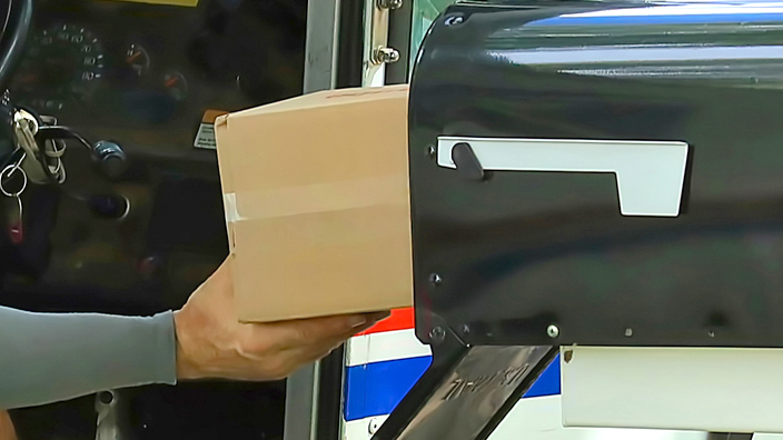 Letter carrier in a delivery vehicle placing a package inside of a black mailbox