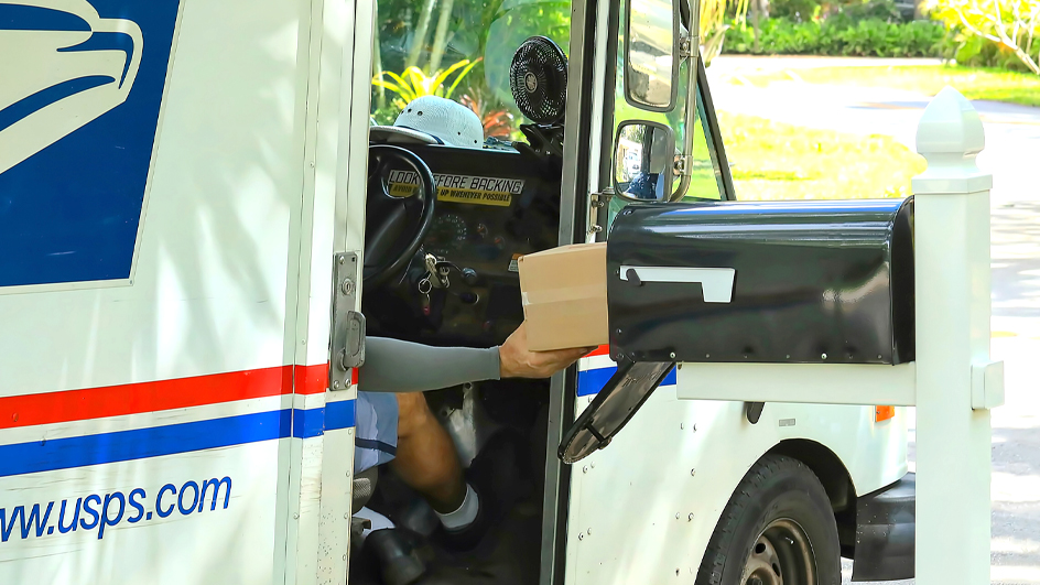 Letter carrier in a delivery vehicle placing a package inside of a black mailbox