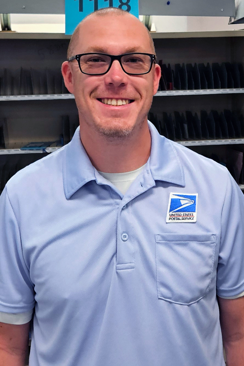 USPS employee wearing glasses and a light blue polo shirt and standing in a mail sorting area