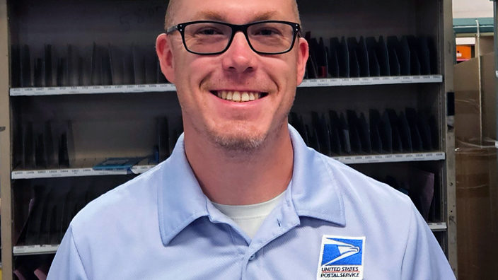 USPS employee wearing glasses and a light blue polo shirt and standing in a mail sorting area
