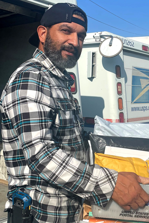 Man standing outside at the back of a USPS delivery vehicle holding a container filled with mail