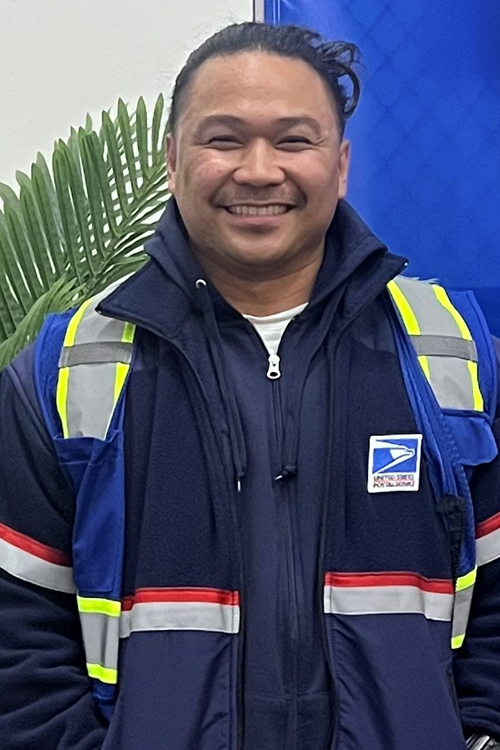 USPS employee wearing a blue jacket with reflective stripes and standing in front of a model USPS tractor-trailer vehicle and a potted plant