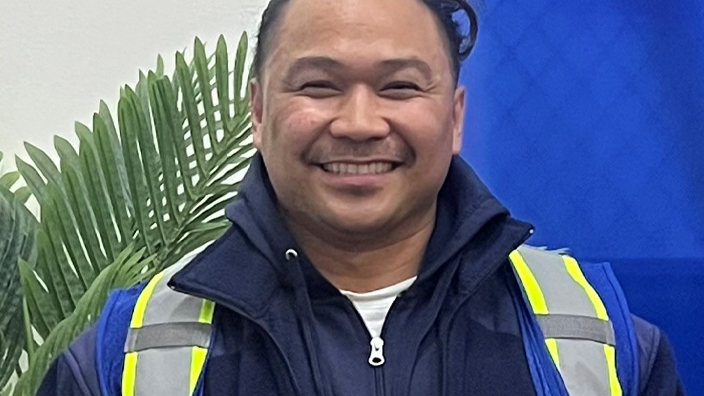 USPS employee wearing a blue jacket with reflective stripes and standing in front of a model USPS tractor-trailer vehicle and a potted plant