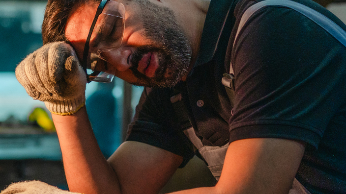 A worker sitting on a stool and wiping sweat from his brow with the back of his hand