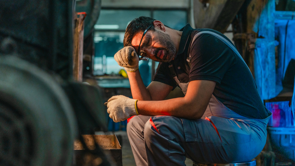A worker sitting on a stool and wiping sweat from his brow with the back of his hand