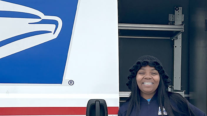 USPS employee in uniform stands outside of a white delivery van