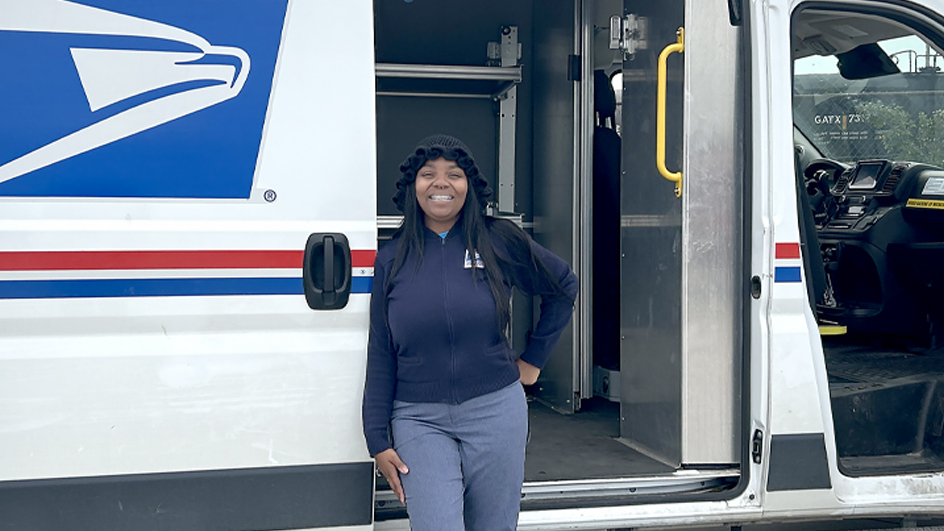 USPS employee in uniform stands outside of a white delivery van