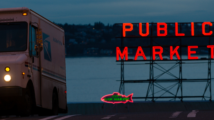 A USPS delivery vehicle driving down the street in darkness away from neon letters spelling out “Public Market”