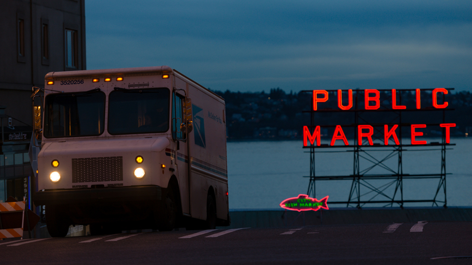 A USPS delivery vehicle driving down the street in darkness away from neon letters spelling out “Public Market”