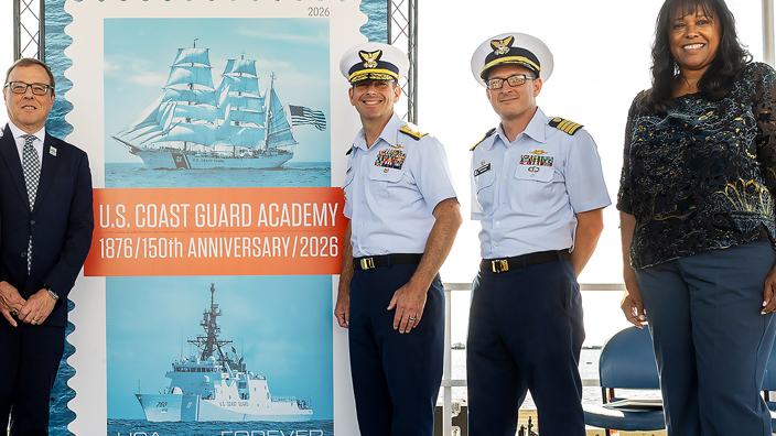 Six people standing on stage beside an enlarged image of the U.S. Coast Guard Academy stamp