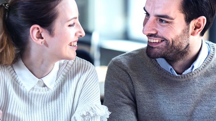 Two co-workers sitting next to each other at a table and looking at one another
