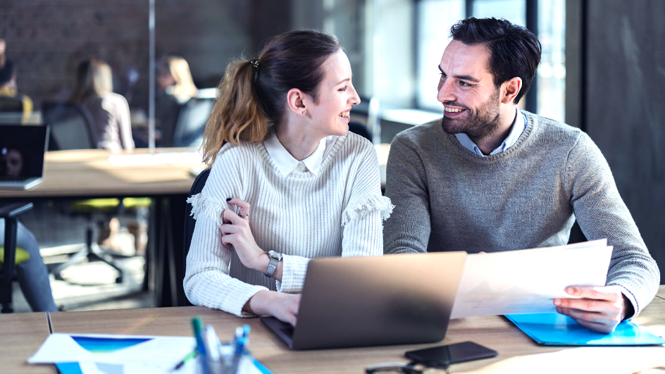 Two co-workers sitting next to each other at a table and looking at one another