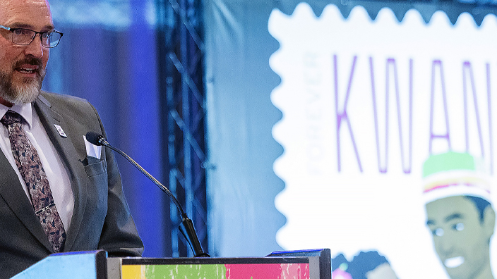 A man in a dark suit standing on stage behind a podium with an enlarged image of the USPS Kwanzaa stamp in the background