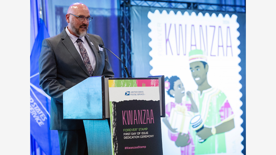 A man in a dark suit standing on stage behind a podium with an enlarged image of the USPS Kwanzaa stamp in the background