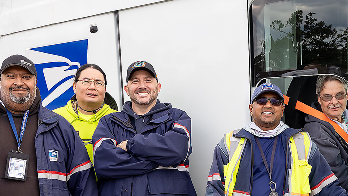 Four USPS employees standing outside of a next-generation delivery vehicle while one employee sits in the vehicle’s driver's seat