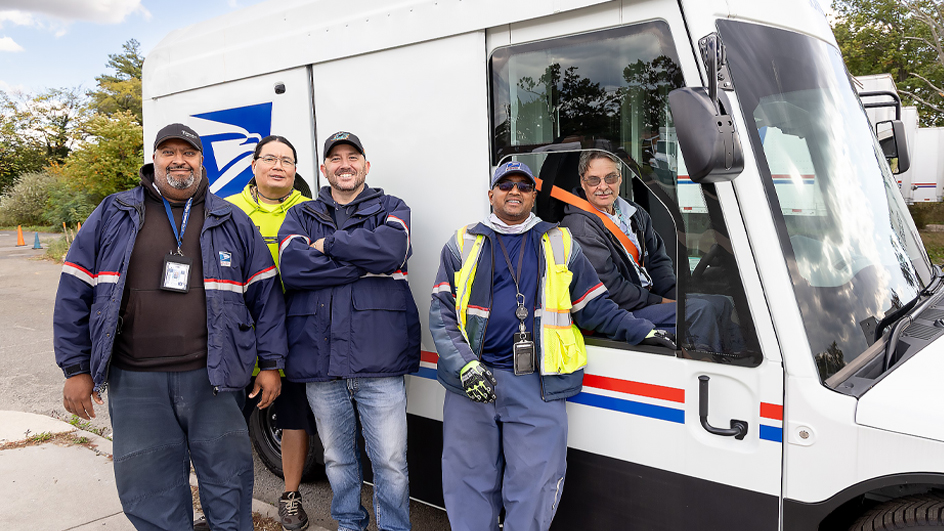 Four USPS employees standing outside of a next-generation delivery vehicle while one employee sits in the vehicle’s driver's seat