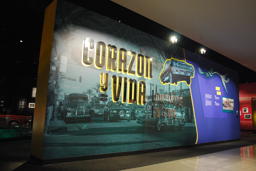 A wall lit from above with a photograph backdrop showing lowriders and a backlit sign reading “Corazón y vida: Lowriding Culture”