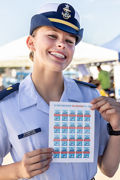 A smiling U.S. Coast Guard Academy cadet in uniform holds up a sheet of U.S. Coast Guard Academy stamps