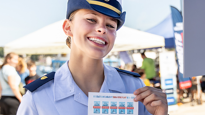 A smiling U.S. Coast Guard Academy cadet in uniform holds up a sheet of U.S. Coast Guard Academy stamps