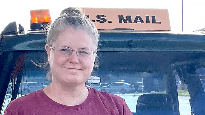 Woman in plum-colored shirt standing outdoors at the rear of a vehicle and next to a tire cover that reads “U.S. rural mail carrier”