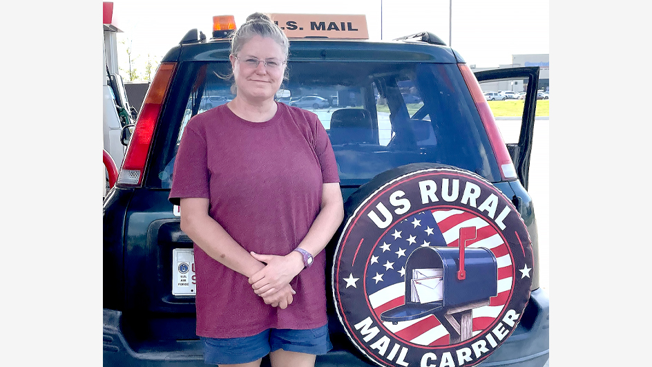 Woman in plum-colored shirt standing outdoors at the rear of a vehicle and next to a tire cover that reads “U.S. rural mail carrier”