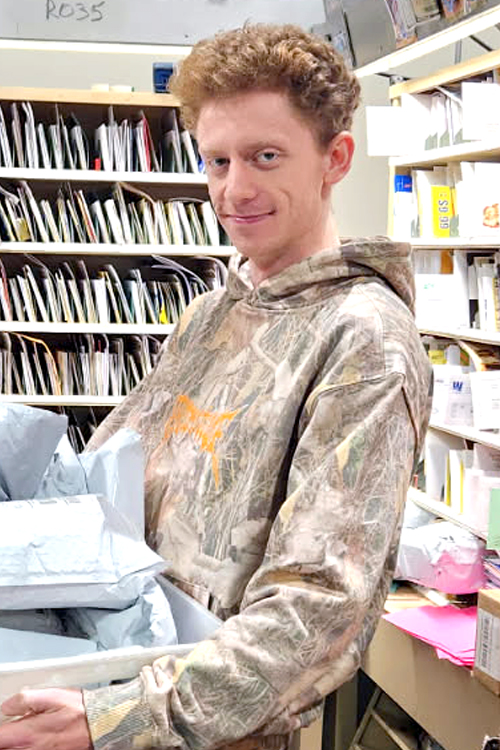 USPS employee wearing a camouflage-patterned sweatshirt and standing in front of rows of sorted mail
