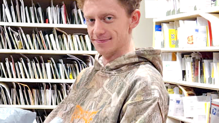 USPS employee wearing a camouflage-patterned sweatshirt and standing in front of rows of sorted mail