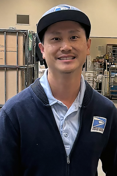 USPS employee in dark blue sweater standing behind a large mail bin on a workroom floor