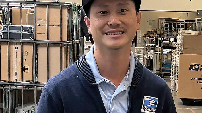 USPS employee in dark blue sweater standing behind a large mail bin on a workroom floor