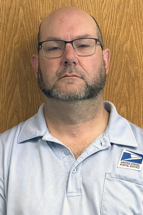 USPS employee wearing a light blue polo shirt and standing in front of a wooden door