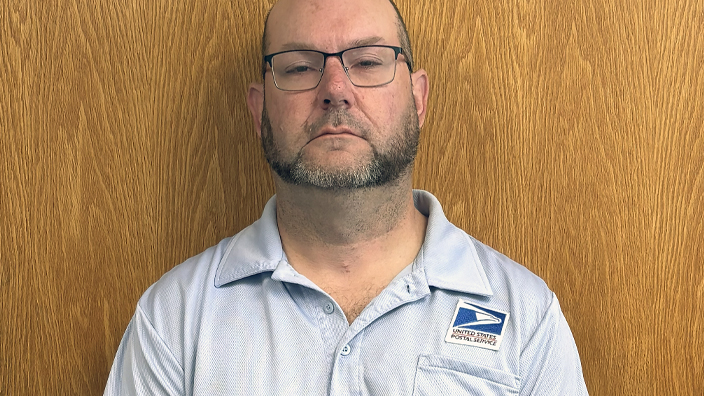 USPS employee wearing a light blue polo shirt and standing in front of a wooden door