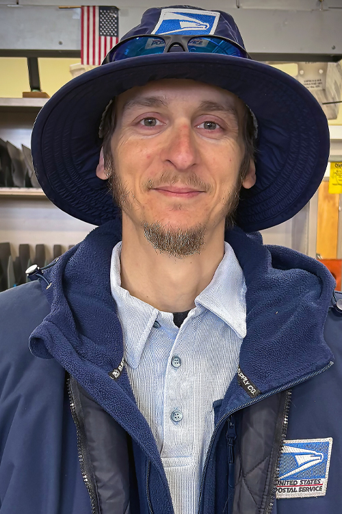 USPS employee wearing a cold-weather postal uniform and standing in a work area