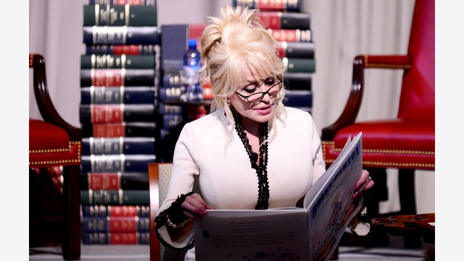 Woman wearing white reading to children from an open book while sitting in front of stacks of books on stage