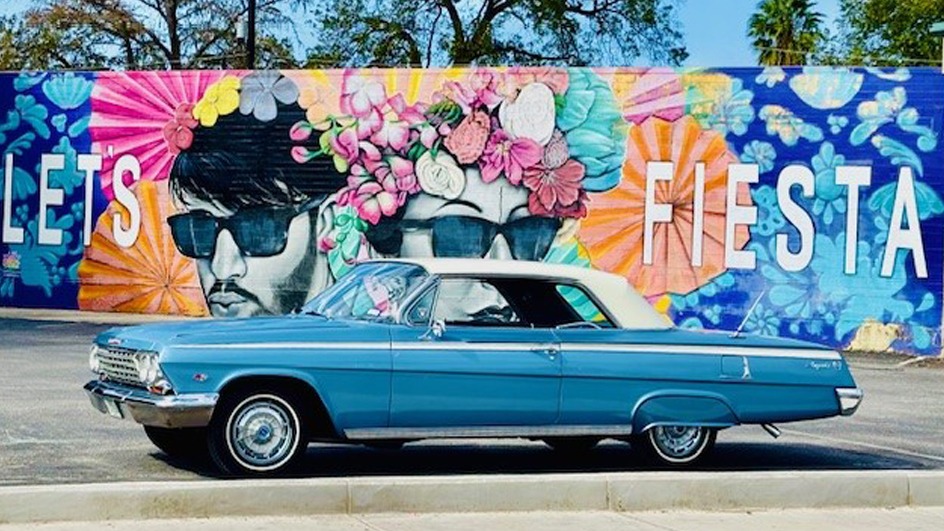 A blue lowrider vehicle parked in front of a colorful mural.