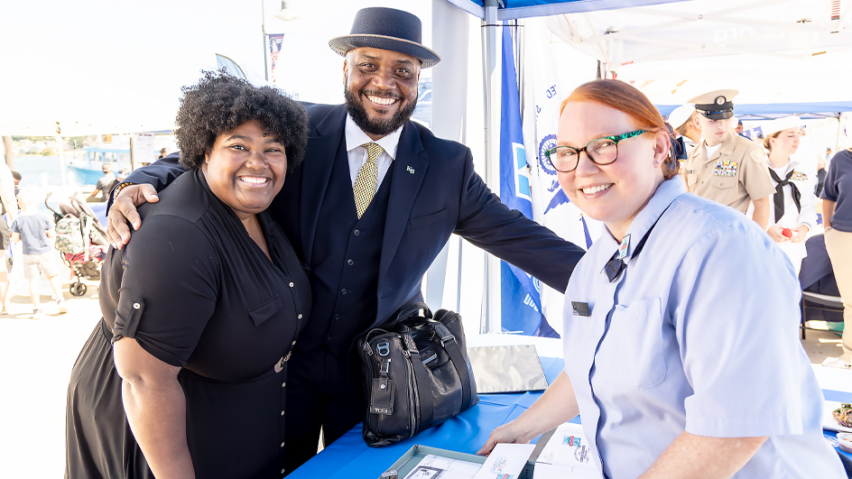 Two adults wearing professional, dark-colored clothing standing next to a uniformed USPS employee with a box of philatelic materials sitting on a table between them