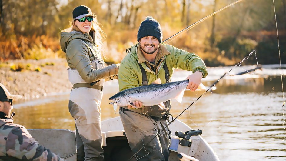 Man holding a large salmon while standing next to a woman holding a fishing road inside of a boat on water
