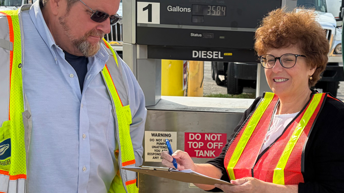 A man and a woman wearing bright-colored safety vests standing side-by-side in front of a fuel pump
